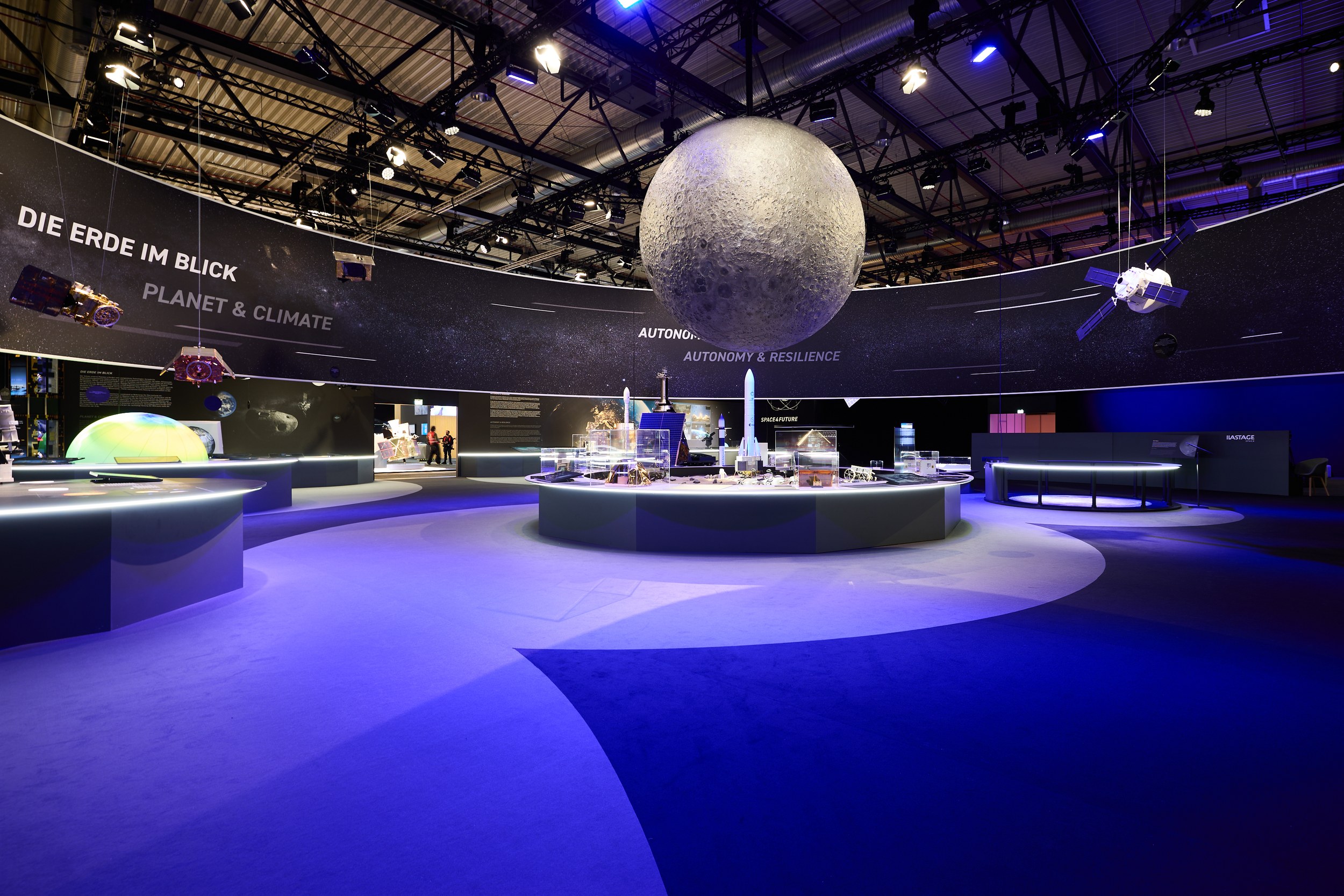 A wide-angle view shows the blue-lit Space Pavilion at ILA Berlin 2026 with a large suspended moon model above the centr