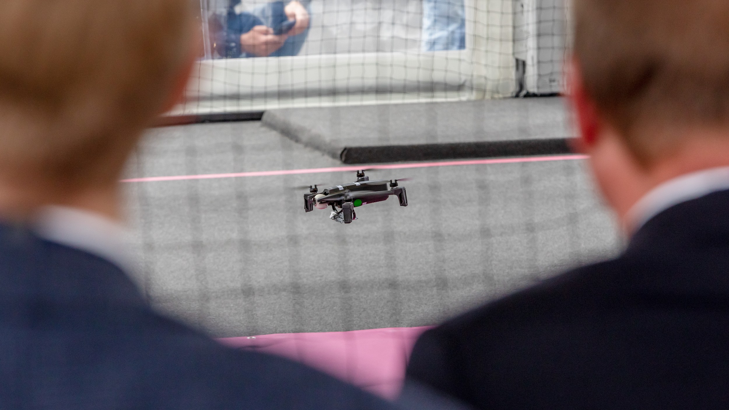A small quadcopter hovers just above the floor inside a flight area enclosed by safety netting. Two large, blurred spect