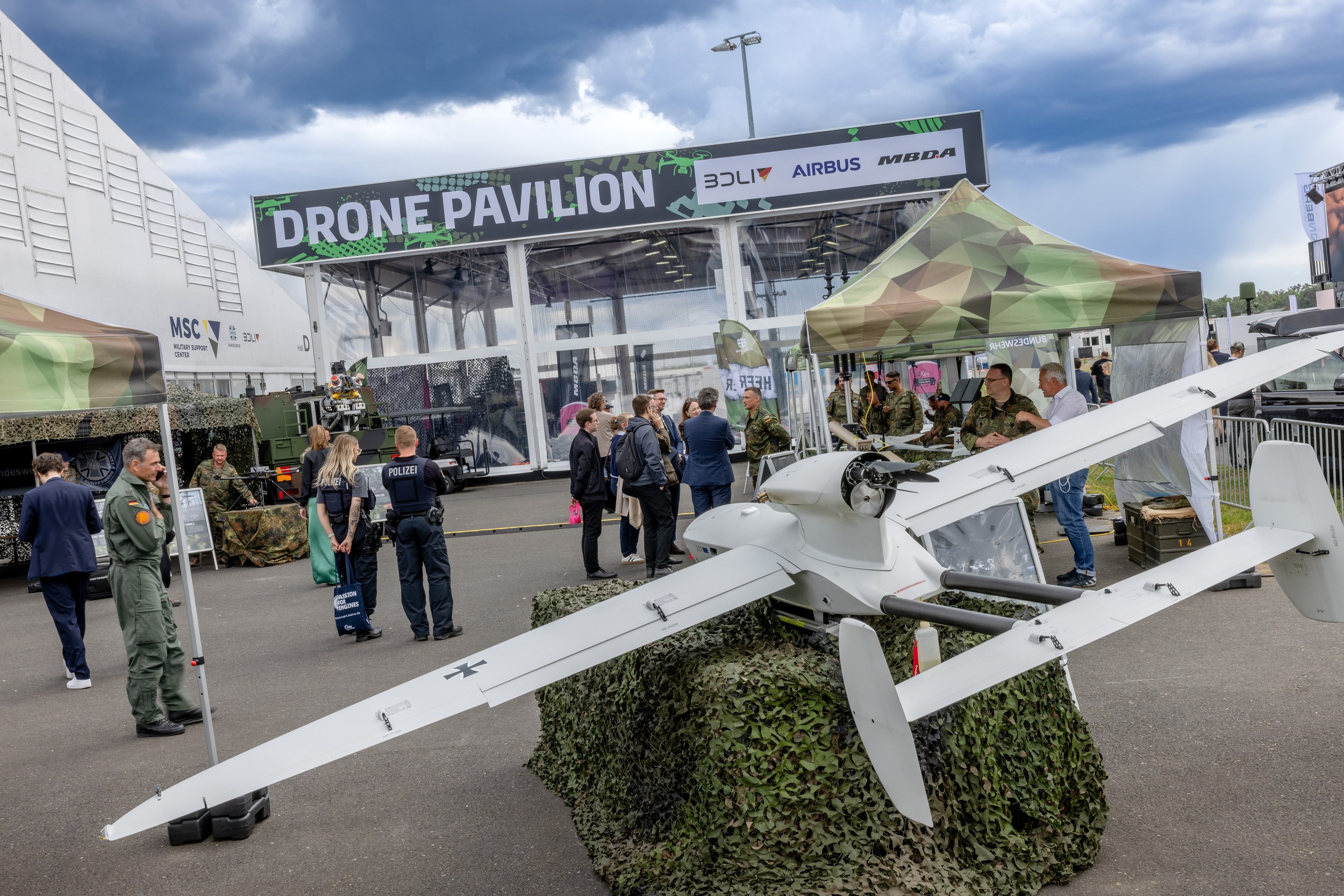 A large military fixed-wing drone is displayed on a camouflage-netted pedestal outside the Drone Pavilion at ILA Berlin 2026. A black Bundeswehr cross is visible on the wing, while visitors speak with uniformed Bundeswehr personnel behind it and two police officers stand to the left of the exhibit.