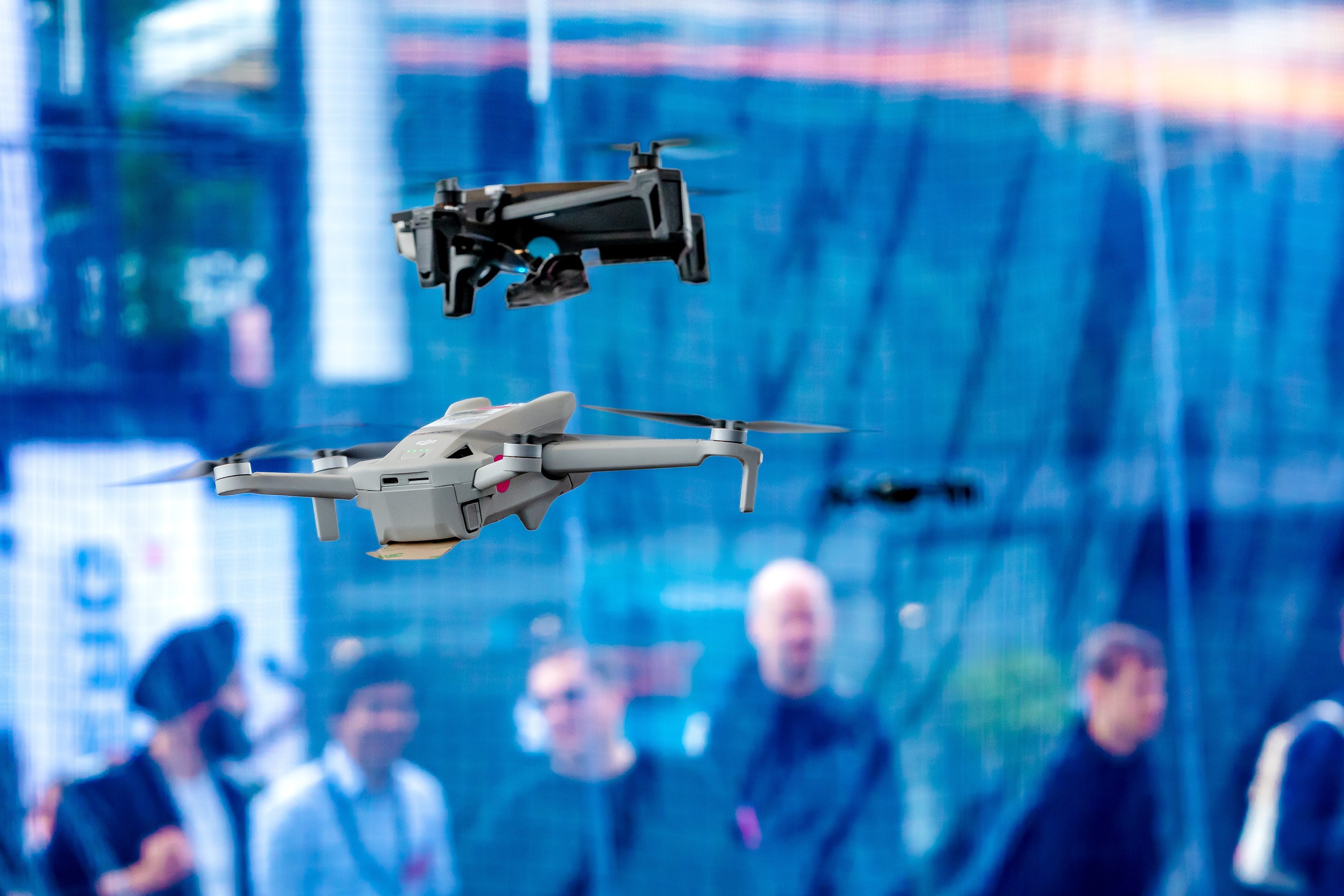 Two clearly visible quadcopters hover one above the other against a blue glazed hall backdrop, with a grey drone in fron