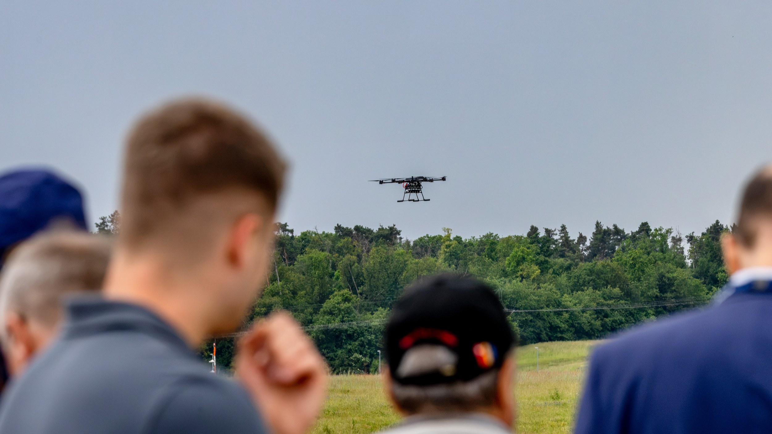 Several blurred and cropped spectators watch a multirotor drone during the Flying Display at ILA Berlin 2026. The open-f