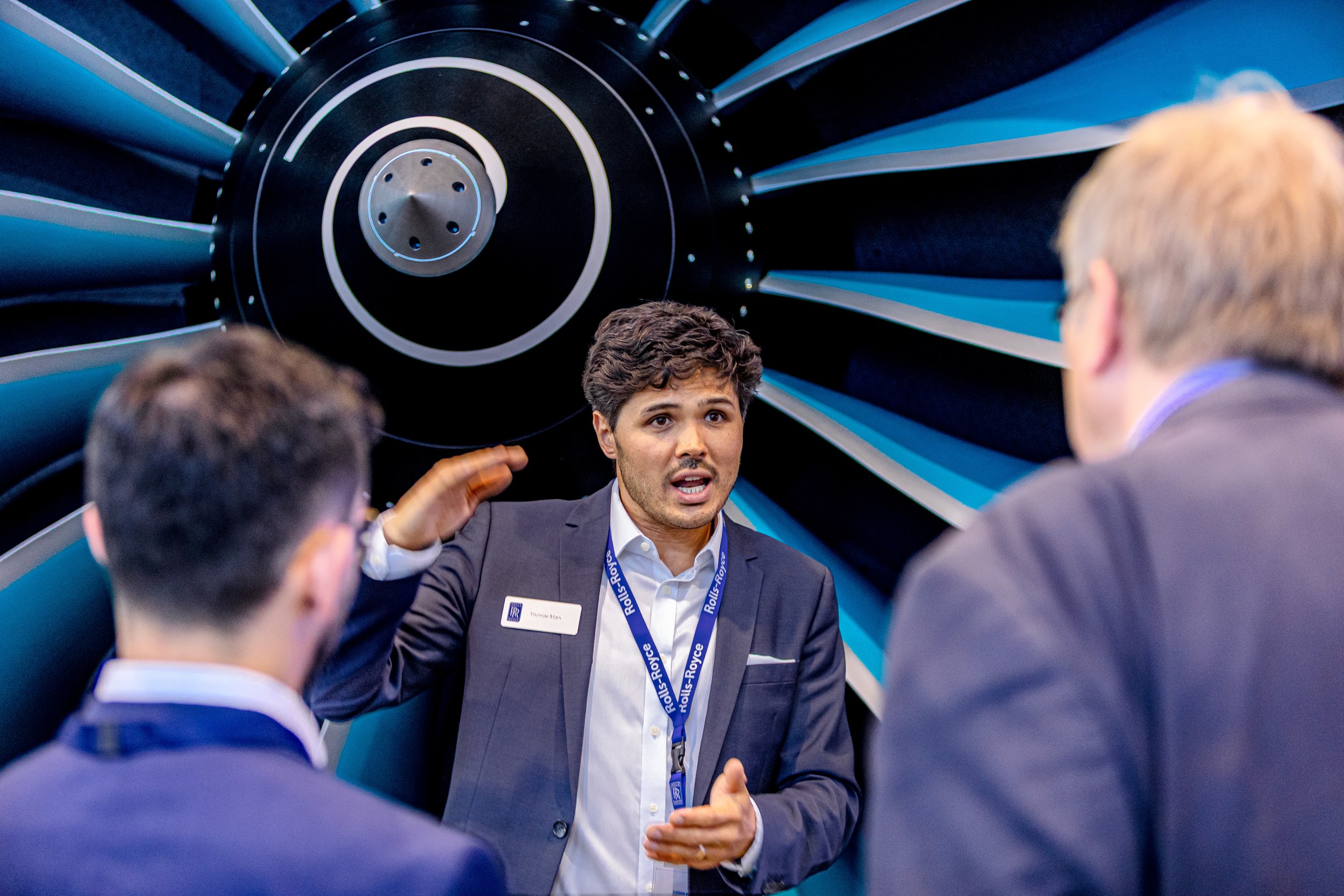 A Rolls-Royce representative explains a technical detail to two visitors in front of a large blue and black turbofan …