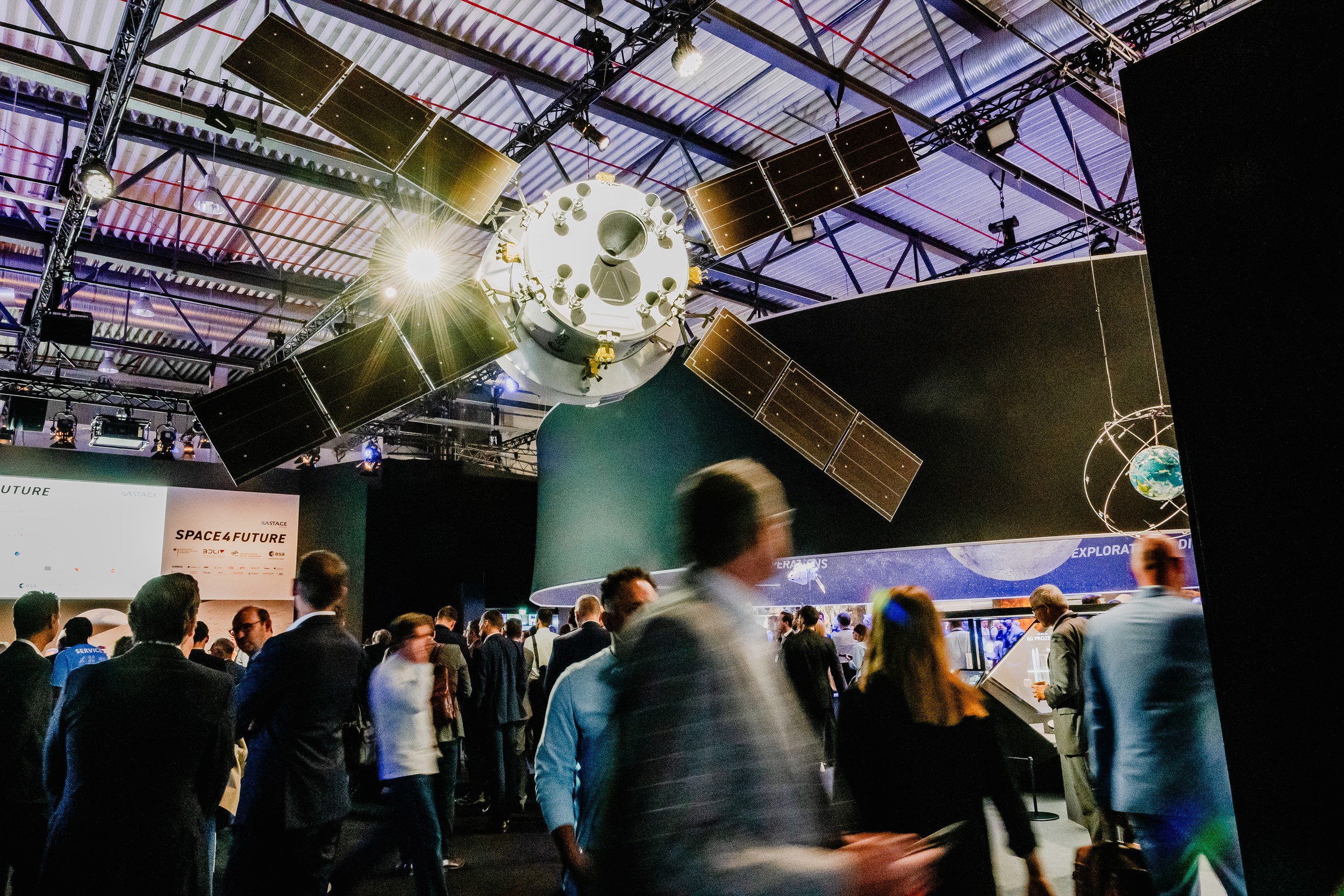 A large space capsule model with four deployed solar panels hangs face-on above a dense crowd in the Space Pavilion at ILA Berlin 2026. Several visitors cross the foreground with motion blur, while the dark exhibition architecture and SPACE4FUTURE branding remain visible behind them.