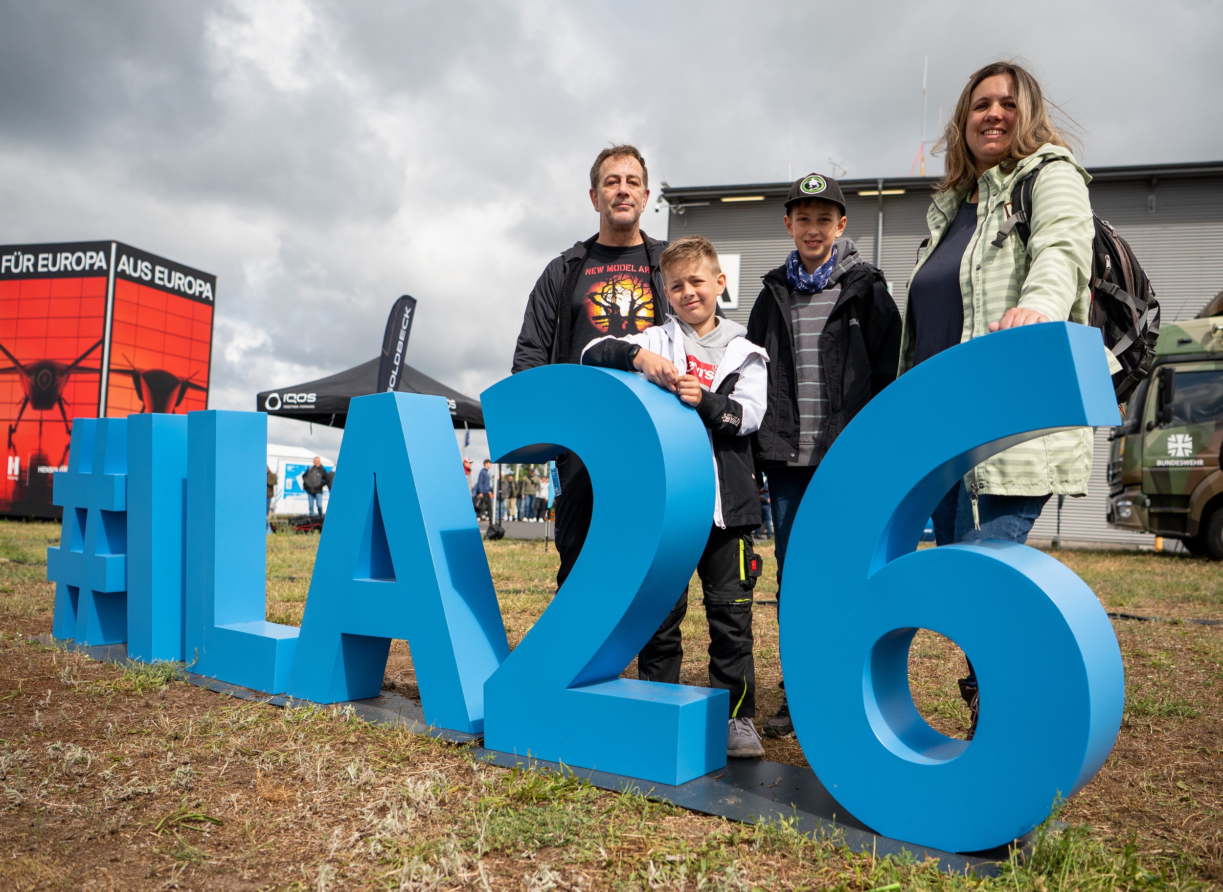 Two adults and two children pose behind the large blue #ILA26 letters in the outdoor area for a souvenir photo.