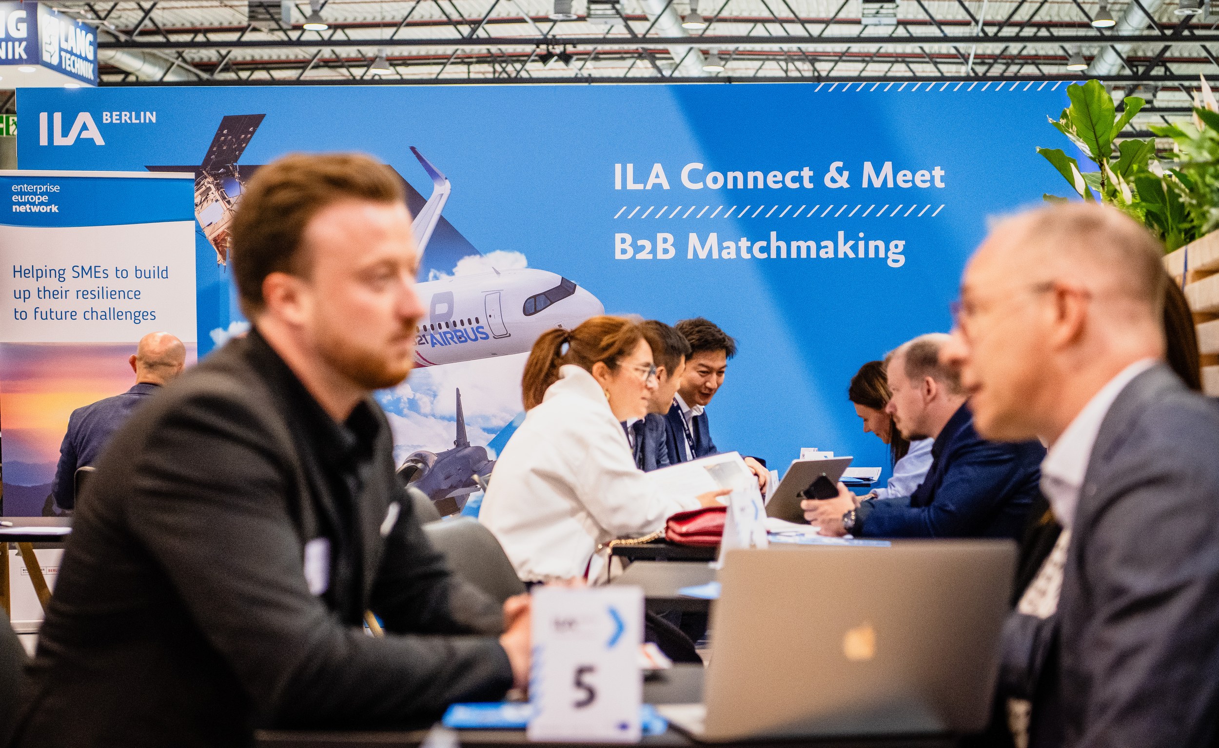 Several pairs hold meetings at numbered tables in front of the large ILA Connect & Meet wall, some working with laptops and tablets. Two men frame the scene in blur in the foreground, with an Enterprise Europe Network banner on the left.