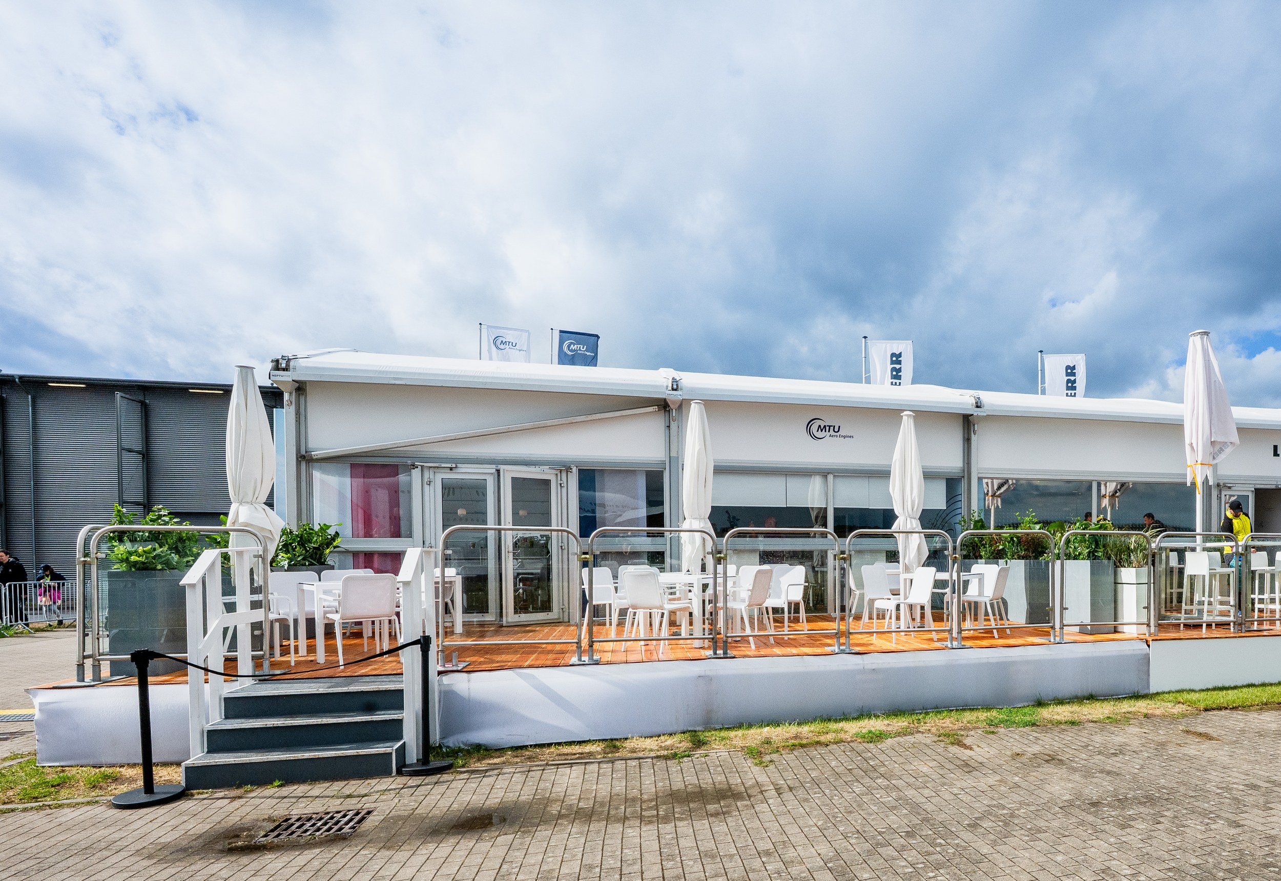 Chalets der ILA 2026 von der Terrassenseite: weiße Module mit Glasfront und zweiflügeliger Glastür, Terrasse mit Glasgeländer und Treppe.