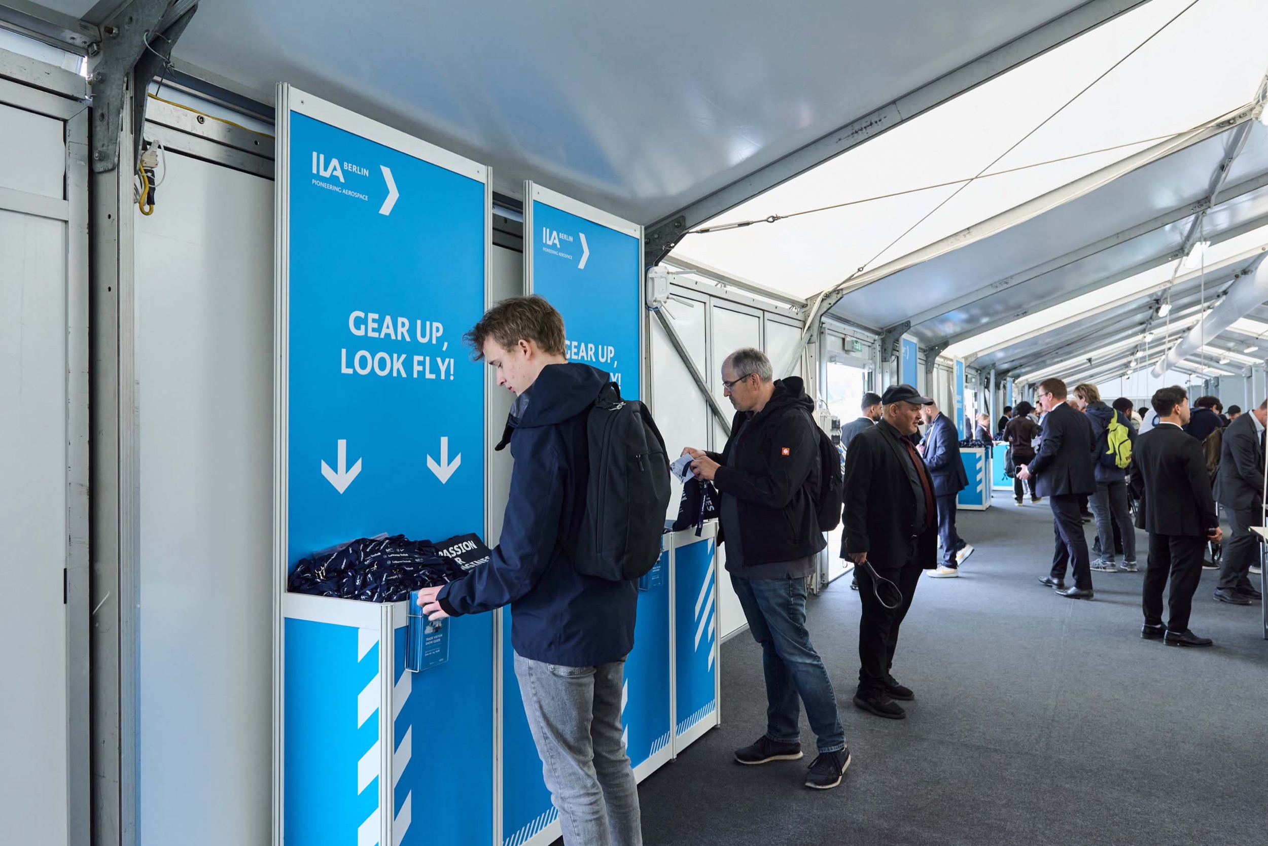 Visitors pick up branded lanyards from the blue gear-up-look-fly dispensers at the ILA 2026 entrance.